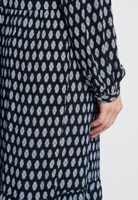 Czarna tkanina sukienki z białymi owalnymi wzorami. Posiada długie rękawy i marszczenia na ramionach. Dołu ma falbaniaste wykończenie.