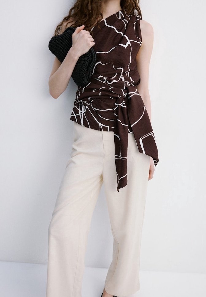 Femme portant un haut asymétrique marron avec des lignes blanches abstraites et un pantalon large beige, tenant une pochette noire texturée.