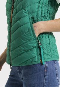 Veste matelassée verte avec une fermeture éclair à l'avant et des poches latérales. Le tissu est lisse avec une finition durable, présentant un design respirant et des accessoires contrastants.
