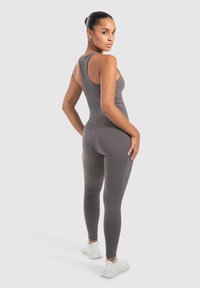 Grå atletisk jumpsuit med en tætsiddende design og uden ærmer. Glat tekstur, accentueret med et minimalistisk logo på ryggen. Hvide sneakers.