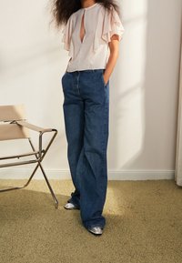 Bluzka w jasnoróżowym kolorze z warstwowymi rękawami z falbanami i dekoltem w kształcie łezki, w połączeniu z niebieskimi dżinsami z szerokimi nogawkami i srebrnymi butami na szpilkach.