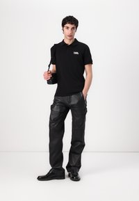 Zwarte poloshirt met logo, gecombineerd met zwarte leren cargobroek met meerdere zakken. Accessoires zijn een kleine zwarte tas.