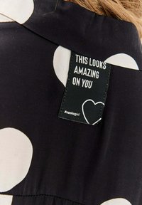 Czarna tkanina z dużymi kremowymi kropkami. Metka z napisem "ŚWIETNIE W TYM WYGLĄDASZ" z graficznym sercem. Tekstura wydaje się gładka.