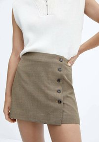 Jupe à carreaux marron avec une fermeture à bouton sur le côté et une longueur courte, associée à un haut sans manches blanc avec un col zippé.