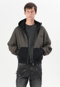 Manga longa oversized preta e verde-oliva com fecho éclair, com um bolso de contraste preto, usada sobre uma camisa cinza, combinada com jeans pretos gastos.
