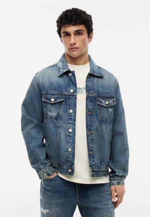 Junger Mann trägt eine ausgewaschene blaue Jeansjacke über einem weißen T-Shirt mit Motiv und passenden ausgewaschenen blauen Jeans, steht mit einer Hand in der Tasche.