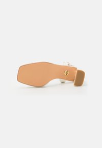 Vit klack sandalett med rund tå och ankelrem. Texturerad tan-soffa med en cirkulär guldslogodetalj. Måttlig klackhöjd.
