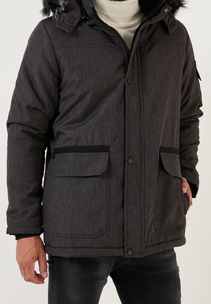 Hombre con abrigo de invierno gris oscuro con capucha forrada de piel, cierres de cremallera y botones a presión, bolsillos con solapa y suéter de cuello alto de color claro.