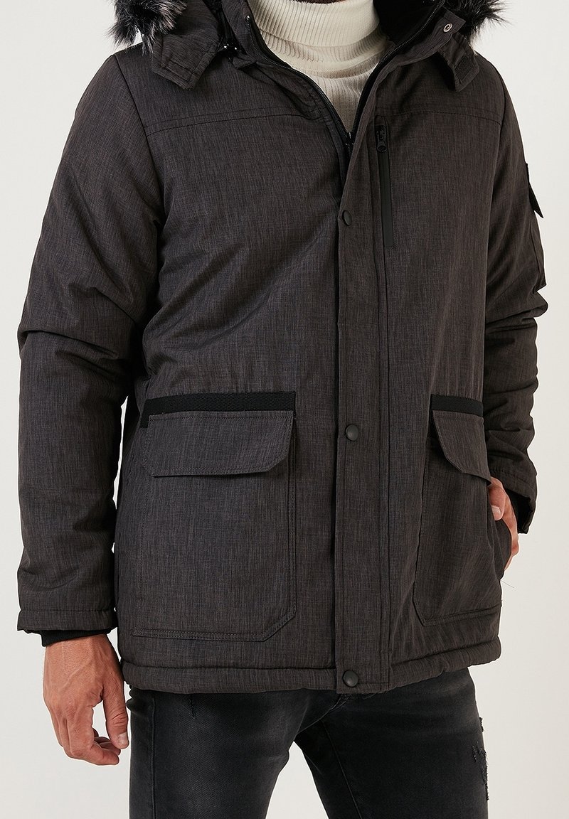 Hombre con abrigo de invierno gris oscuro con capucha forrada de piel, cierres de cremallera y botones a presión, bolsillos con solapa y suéter de cuello alto de color claro.