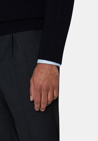 Pull en laine navy avec des poignets côtelés, superposé à une chemise à rayures bleu clair, porté avec un pantalon tailleur gris foncé et une main visible.