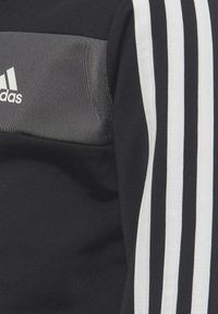 Haut de sport noir avec un panneau gris et trois bandes blanches le long des manches. Fabriqué en tissu texturé avec un logo Adidas.