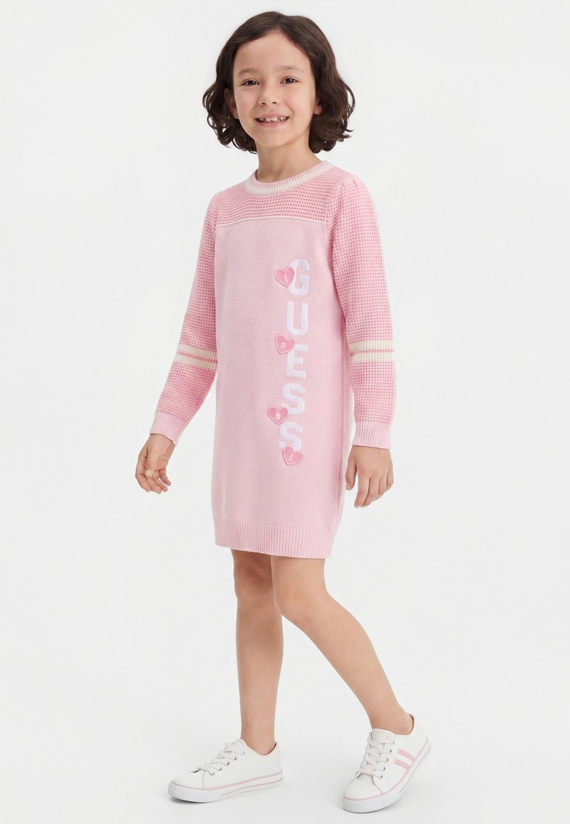 Jeune fille souriante, vêtue d'une robe-pull rose à manches longues avec l'inscription « GUESS » et des cœurs, associée à des baskets blanches, debout sur fond blanc.