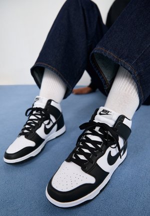Personne portant des baskets montantes Nike noir et blanc avec des chaussettes blanches et un jean bleu foncé assise sur un tapis bleu.
