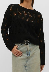 Zwarte gebreide trui met opengewerkte structuur, voorzien van lange mouwen en een geribde halslijn, gecombineerd met een lichte denim rok.