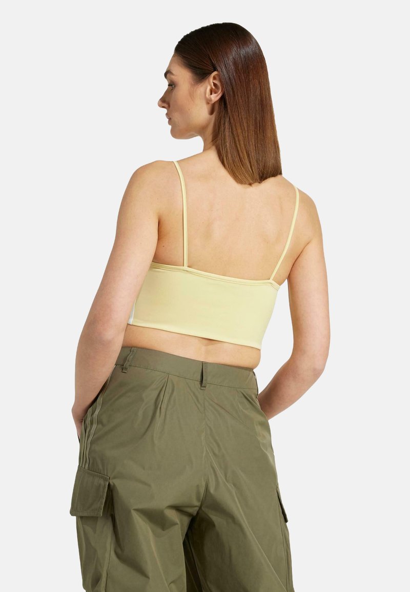 Femme aux cheveux bruns raides portant un crop top jaune à fines bretelles et un pantalon cargo vert olive, se tenant les mains dans les poches.