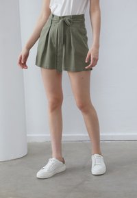 Olivgrüne, hoch taillierte Shorts mit Falten, Bindeband an der Taille und glatter Textur. Kombiniert mit weißen Sneakers.