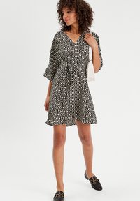 Robe à motif ondulé noir et blanc avec un décolleté en V, des manches courtes évasées, une taille cintrée et une jupe fluide. Associée à des mocassins marron et un sac.