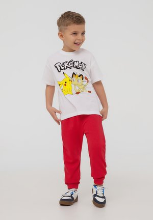 Biała koszulka z grafiką Pokémon, na której znajdują się Pikachu i Meowth, w zestawie z jasnoczerwonymi spodniami dresowymi i granatowymi trampkami.