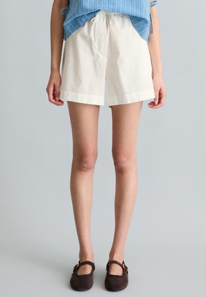 Kobieta w białych szortach ze ściągaczem, niebieskim topie bez rękawów oraz ciemnobrązowych butach Mary Jane, stojąca na jednolitym tle.