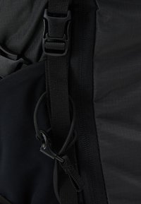 Černá nylonová taška s nastavitelnými popruhy, uzávěrem na klip a elastickým detailem. Texturovaný povrch s hladkými akcenty.