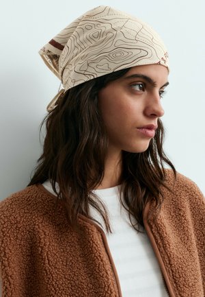 Jeune femme aux longs cheveux foncés portant un foulard beige clair à motifs, une veste marron texturée et une chemise blanche, regardant vers la droite.
