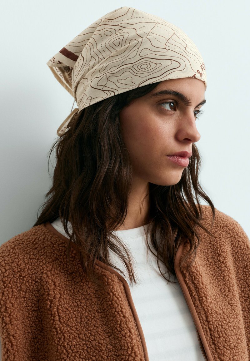 Jeune femme aux longs cheveux foncés portant un foulard beige clair à motifs, une veste marron texturée et une chemise blanche, regardant vers la droite.