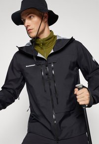 Chaqueta negra impermeable con acentos grises, dos bolsillos en el pecho con cremallera, capucha ajustable y un acabado texturizado. El modelo sostiene un bastón.