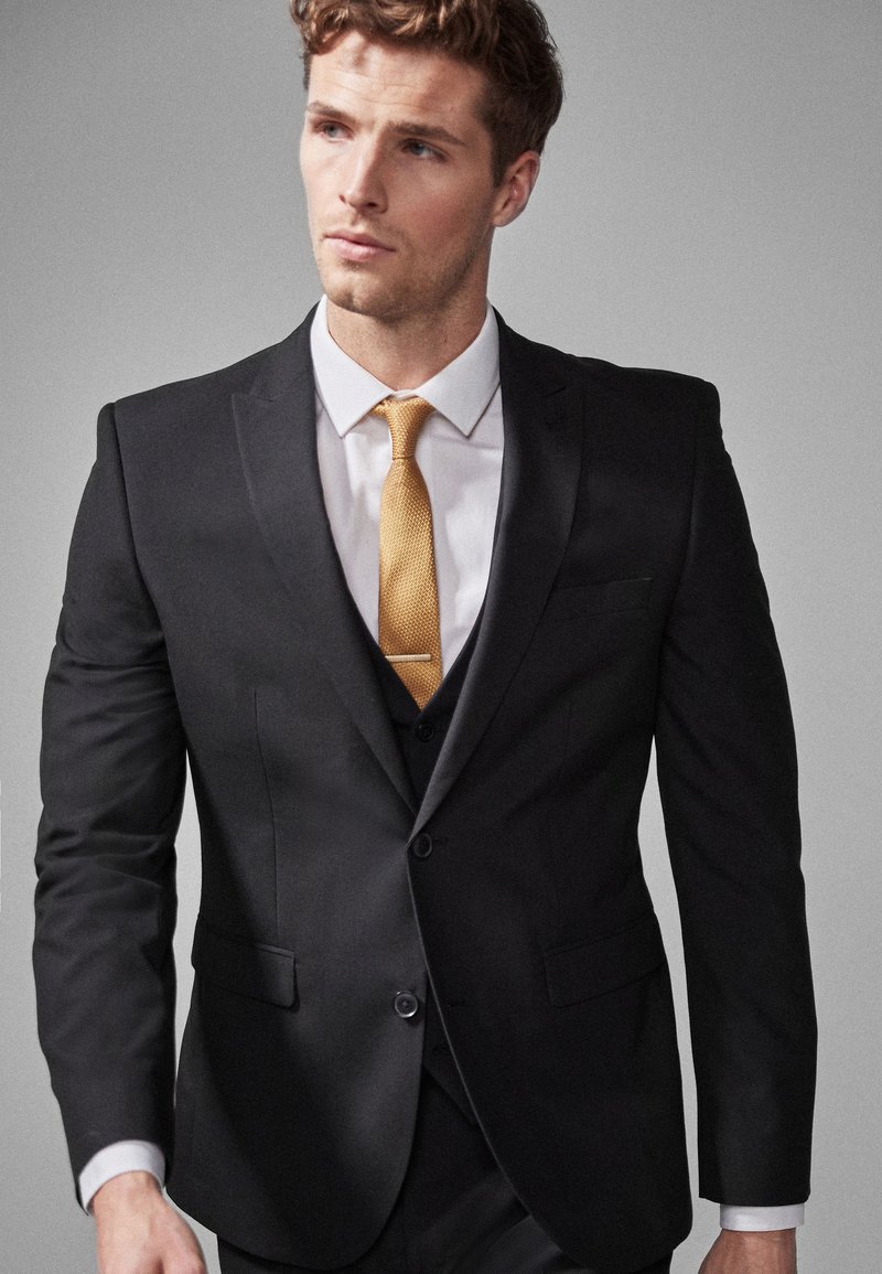 Black suit with matching lapels, white shirt, and a textured mustard tie. Notable details include a slim fit and tailored stitching.