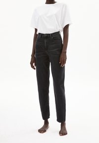 Jeans negros de cintura alta con corte recto, hechos de denim, combinados con una camiseta de algodón blanca de corte holgado. Modelo descalzo.