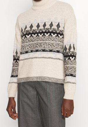 Beige tyndhals sweater med sorte og grå geometriske mønstre, båret med grå teksturerede bukser. Hænder afslappet langs siderne.