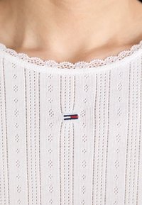 Hvid top med horisontale striber og detaljeret eyelet-design. Scallop-lace udskæring med et lille broderet logo i rød og blå.