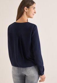Marineblå langærmet bluse med struktureret overflade, rund halsudskæring og en central rygfold, parret med lysegrå slidte jeans.