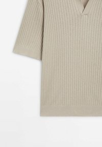 Beige ribbipaita lyhyillä hihnoilla ja V-pääntiellä. Ominaisuuksiin kuuluu pehmeä tekstuuri ja suora helma, joka esittää pystysuoria viivoja kauttaaltaan.