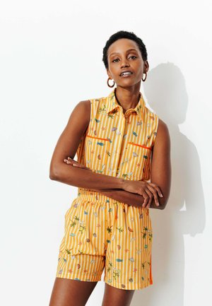 Femme portant une combinaison sans manches jaune rayé avec des motifs colorés de fruits et de feuilles, debout, les bras croisés, sur un fond blanc.