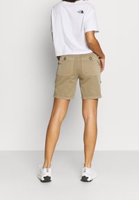 Person som bär beige cargoshorts, vit cropped t-shirt från The North Face och vita sneakers, stående vänd bort från kameran mot en enfärgad bakgrund.