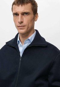 Giacca blu navy con zip e collo alto, realizzata in tessuto texturizzato. Indossata sopra una camicia azzurra a righe, con chiusura a zip liscia.