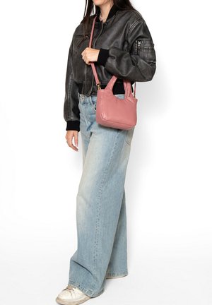 Vrouw die een zwarte leren bomberjack draagt, lichtblauwe jeans met wijde pijpen, witte sneakers, en een kleine roze handtas over haar schouder draagt.