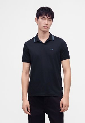 Jeune homme aux cheveux courts noirs portant un polo noir à manches courtes avec des accents bleus, debout devant un fond clair uni.