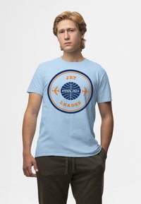 Maglietta in cotone azzurro chiaro con un design circolare che presenta le scritte "JET" e "LEADER" in arancione e un logo "PAN AM" blu navy con grafica di aerei.