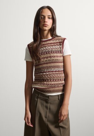 Junge Frau mit langen dunklen Haaren, die eine gemusterte Strickweste über einem weißen kurzärmligen Hemd und olivgrünen weiten Hosen trägt.