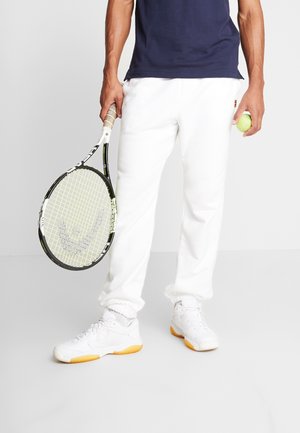 Homme portant un pantalon et des chaussures de tennis blancs, tenant une raquette de tennis dans une main et une balle de tennis dans l'autre, sur un fond uni.