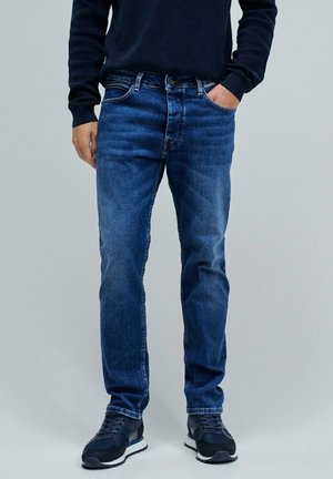 Jeans de mezclilla en azul medio con un ajuste clásico, que cuentan con un cierre de botón y cremallera, cinco bolsillos y un ligero desgaste en las piernas.
