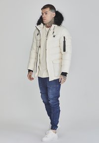 Puffjakke i creme med pelsforet hætte, frontlommer og elastiske manchetter, parret med marineblå jeans og hvide sneakers.