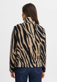 Veste en polaire avec un motif zèbre beige et noir, col montant et tissu texturé. Coupe décontractée, portée avec un jean sombre.