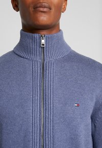 Blå zip-up tröja i ett fint stickat mönster, med en ribbad hög krage, subtila vertikala ränder och en liten logotyp på vänster bröst.