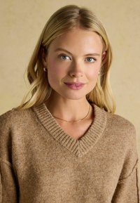 Beżowy sweter w serek, wykonany z teksturowanego dzianiny, z ribbowanym dekoltem i luźnym krojem. Towarzyszy mu delikatny złoty naszyjnik.