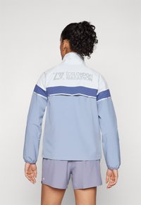 Giacca azzurra con accenti bianchi e blu navy, colletto alto e una stampa grafica sulla schiena. Pantaloni corti viola completano l'outfit.