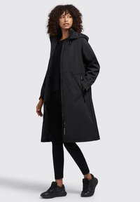 Parka nera impermeabile con cappuccio, zip intera, tasche laterali e silhouette lunga, indossata sopra abbigliamento nero e scarpe da ginnastica.