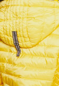 Veste matelassée jaune avec une boucle en tissu rayée marron et noire attachée à l'arrière près du col.