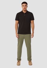 Zwarte polo met twee knopen, gecombineerd met olijfgroene broek. Model draagt beige schoenen. Eenvoudig ontwerp, korte mouwen en platte kraag.